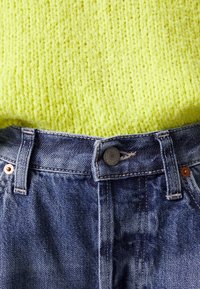 Gros plan sur un jean en denim bleu boutonné à la taille sous un pull jaune vif en maille texturée.