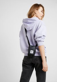 Bolso cruzado negro con un logo blanco sujeto a una correa negra, llevado sobre una sudadera lavanda y jeans oscuros. Tejido suave con un hardware mínimo.
