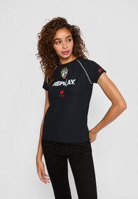 Vrouw met krullend haar, gekleed in een zwart T-shirt van het merk Replay en zwarte broek, staand met één hand op de heup en kijkend opzij.