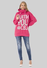 Rosa übergroßer Kapuzenpullover mit weißem Grafiktext, ausgestattet mit einem Cowl-Neck und Kordeln. Kombiniert mit blauen Skinny-Jeans mit Nahtdetails.