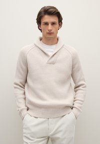 Hellbeiger gerippter Strickpullover mit Schalkragen und V-förmigem Ausschnitt. Über einem weißen Hemd getragen, kombiniert mit hellen Hosen.