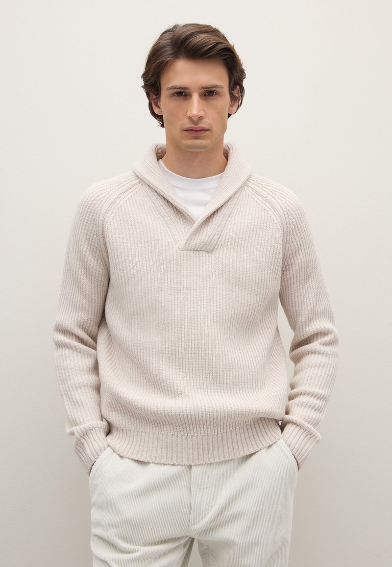 Hellbeiger gerippter Strickpullover mit Schalkragen und V-förmigem Ausschnitt. Über einem weißen Hemd getragen, kombiniert mit hellen Hosen.