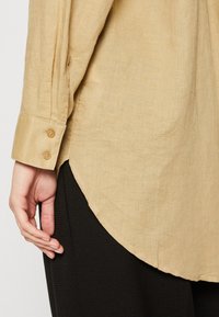 Poignet de chemise en lin beige à manches longues avec deux boutons portée sur un pantalon noir texturé, main détendue sur le côté.