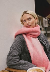 Pink wool scarf draped around the neck, soft texture, gradient color. Gray quilted jacket with visible stitching. Wooden table with coffee cup.