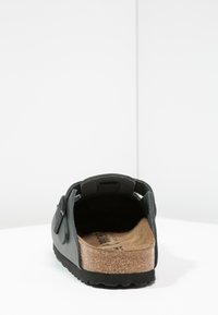 Svarte slip-on sko med kork såle, åpen bakdel og teksturert gummisåle. Har en dekorativ stropp på overdelen.