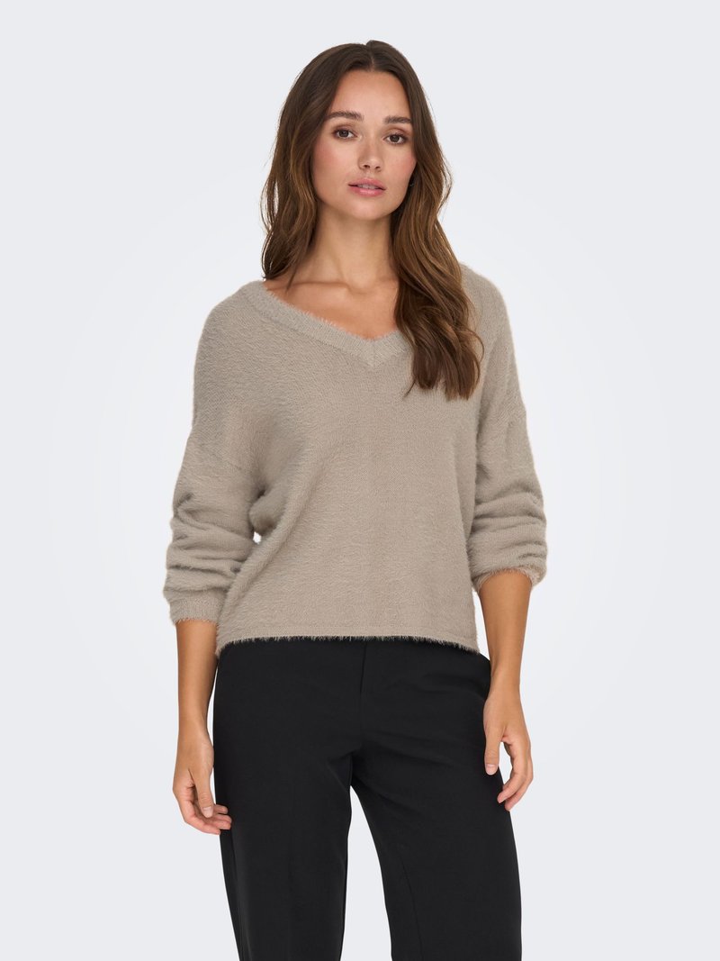 Woman with long brown hair wearing a beige fuzzy V-neck sweater and black pants against a plain light background.