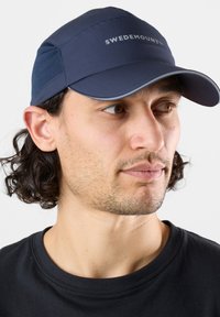 Cap blu navy con visiera curvata, realizzato in tessuto testurizzato. Presenta il logo "SWEDEMOUNT" in grigio chiaro. Indossato con una maglietta nera.
