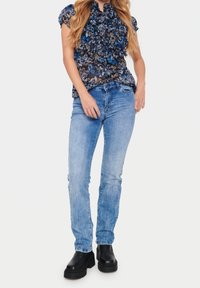 Nainen pukeutuneena siniseen kukkakuosisella röyhelöblusilla, vaaleansinisissä slim fit -farkuissa ja mustissa paksupohjaisissa saappaissa, seisoo tasaisen taustan edessä.