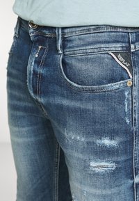 Modré džíny z denimu se slim fit střihem, s efektem opotřebení, jemným blednutím a značkovou nášivkou na zadní kapse. Kovové knoflíky a nýty jako akcenty.