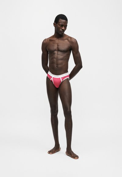 Μυώδης άντρας με σκουρόχρωμο δέρμα που φοράει ροζ σλιπ με λευκή μέση, όρθιος ξυπόλητος μπροστά σε απλό ανοιχτό γκρι φόντο.