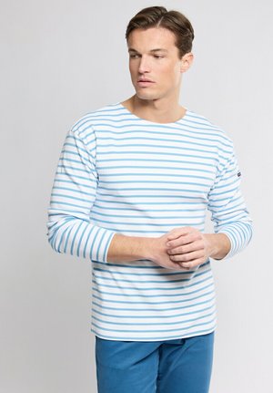Jeune homme aux cheveux bruns portant une chemise à manches longues blanche avec des rayures horizontales bleu clair, mains jointes, regardant sur le côté.
