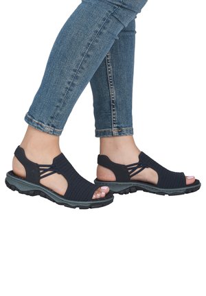 Pieds portant des sandales tissées bleues à bouts ouverts et des jeans bleus retroussés, montrant des chaussures décontractées d’extérieur avec des semelles robustes.