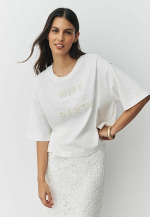 Mosolygó nő fehér ingben, amelyen „WIFE OF THE PARTY” felirat gyöngyszemekkel, és fehér virágos csipkerokával egyszerű háttér előtt.