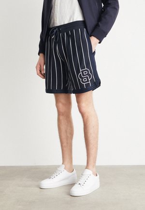 Mann trägt marineblaue Shorts mit feinen Nadelstreifen und weißem, gesticktem "B"-Logo, weiße Sneaker, marineblaue Jacke und weißes Hemd, steht auf einem grauen Boden.