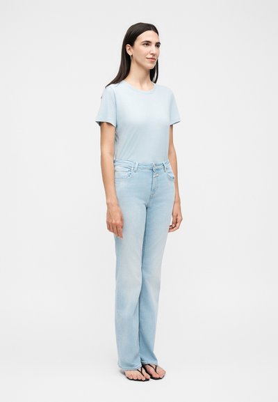 Γυναίκα όρθια, φοράει ανοιχτό μπλε κοντομάνικη μπλούζα, ανοιχτό μπλε τζιν και μαύρες σαγιονάρες σε λευκό φόντο.