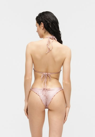 Hosszú sötét hajú nő világos rózsaszín, megkötős bikiniben, háttal áll egy egyszerű világos háttér előtt.
