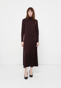 Robe longue ajustée en bordeaux foncé avec un col haut et des manches longues. Texture lisse, longueur cheville avec des fentes latérales pour plus de liberté de mouvement.