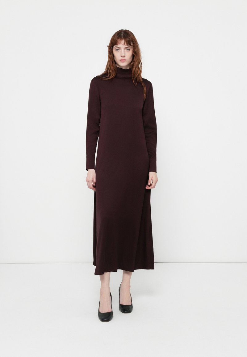 Robe longue ajustée en bordeaux foncé avec un col haut et des manches longues. Texture lisse, longueur cheville avec des fentes latérales pour plus de liberté de mouvement.