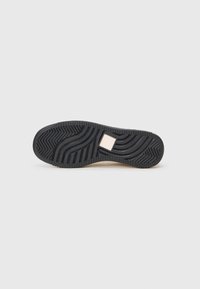 Must musta sole, millel on sakilised ja lainekujulised mustrid, ning keskosas väike ruudukujuline beež silt, valgel taustal.