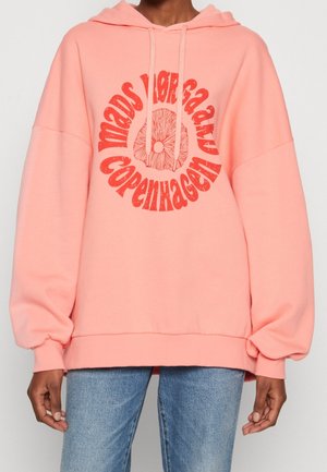 Person klädd i en överdimensionerad rosa hoodie med röd cirkulär text och grafik "mads nørgaard copenhagen", kombinerad med blå jeans.
