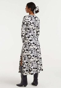 Largo vestido floral en blanco y negro con mangas largas, aberturas laterales y una textura suave, combinado con botas negras hasta la rodilla y una cinta para el cabello de patrón.