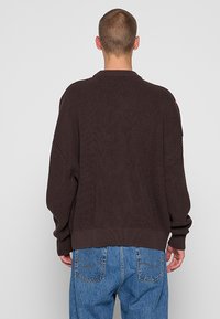 Pull tricoté marron foncé à coupe décontractée, manches larges et ourlet côtelé. Orné d'une petite étiquette logo sur l'épaule.