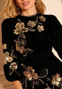 Rochie din catifea neagră cu broderie florală din paiete aurii și nuanțe metalice, cu talie cambrată și mâneci bufante.