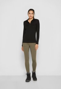 Czarna bluzka z długim rękawem i kołnierzem zapinanym na ćwierć suwak, zestawiona z oliwkowymi legginsami z bocznymi kieszeniami i czarnymi butami na sznurówki.