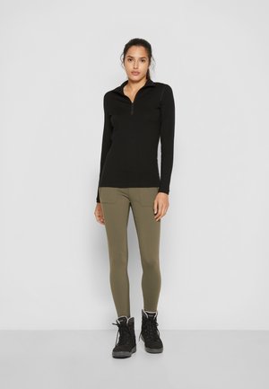 Czarna bluzka z długim rękawem i kołnierzem zapinanym na ćwierć suwak, zestawiona z oliwkowymi legginsami z bocznymi kieszeniami i czarnymi butami na sznurówki.