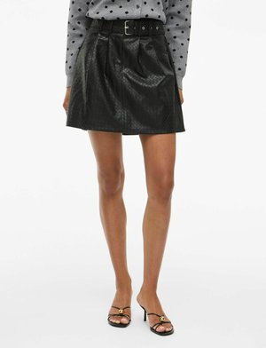 Jupe mini en simili cuir noir avec un motif en losanges texturé, taille ceinturée et plis ; portée avec des sandales à talons noires.
