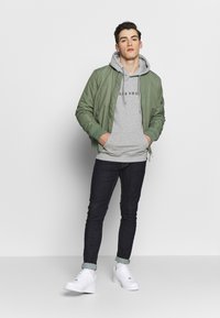 Olijfgroen bomberjack over een grijze hoodie, gecombineerd met donkere jeans en witte sneakers. De outfit heeft een casual pasvorm en textuurcontrasten.