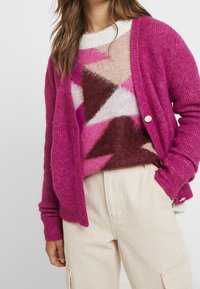 Femme portant un pantalon cargo crème, un pull en laine à motifs géométriques rose, marron et crème, ainsi qu'un cardigan ouvert magenta avec des boutons dorés.
