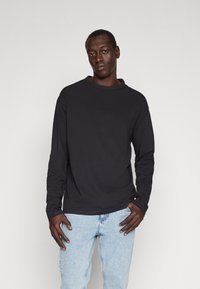 Mand iført en ensfarvet sort langærmet trøje og lyseblå jeans stående mod en ensartet lysegrå baggrund.