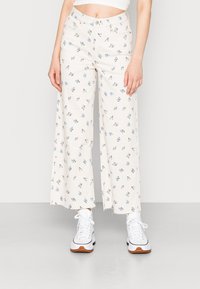 Calças largas florais em creme, com padrões de flores azuis, bainha desfiada, duas bolsas laterais, combinadas com ténis brancos.