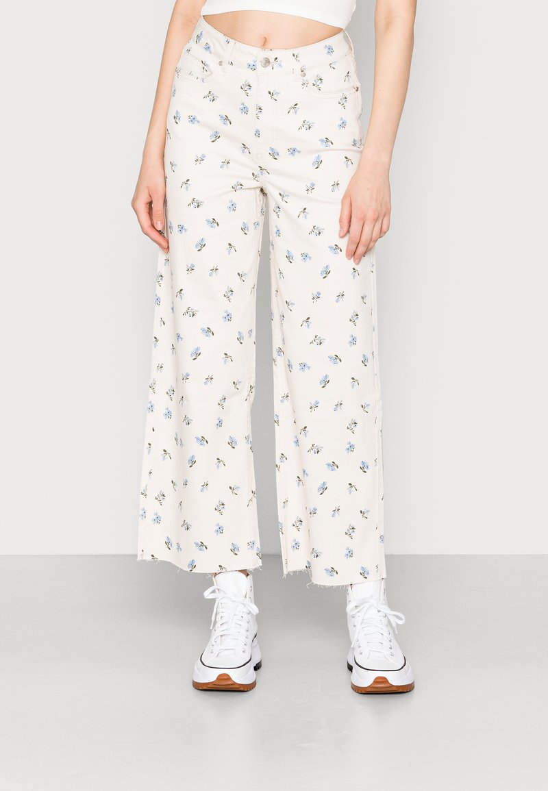 Calças largas florais em creme, com padrões de flores azuis, bainha desfiada, duas bolsas laterais, combinadas com ténis brancos.