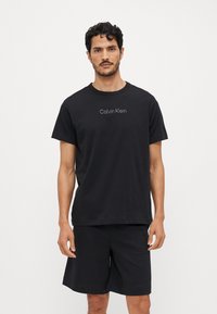 Crna pamučna majica s kratkim rukavima, opuštenog kroja, sa logotipom "Calvin Klein" u svjetlijoj nijansi. Uparena s crnim kratkim hlačama.