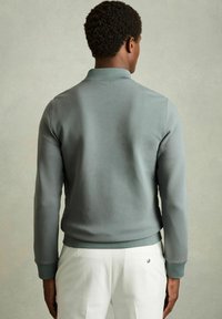 Hanorac gri, material neted, manșete și guler ribanați, design ajustat, văzut din spate, asortat cu pantaloni albi.