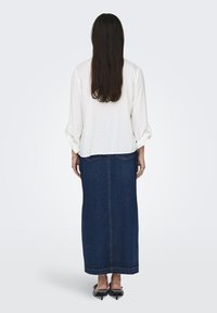 Bluză albă cu buline, cu mânecile suflecate, combinată cu o fustă lungă din denim închis. Ținuta are o siluetă dreaptă și un design simplu.
