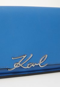 Plava kožna torbica s teksturiranom površinom koja sadrži istaknuti srebrni logo "Karl". Uključuje crne naglaske duž donjeg dijela.