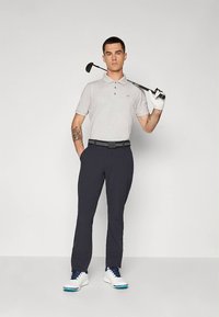 Šviesiai pilka polo marškinėliai su apykakle, tamsiai mėlynos kelnės, juodas diržas, balti golfo batai su mėlynomis detalėmis. Modelis laiko golfo lazdą ant peties.