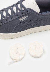 Puma nubuck teniske v temno modri barvi z belimi detajli, z teksturirano zgornjščino, ravnimi vezalkami in kontrastnim gumijastim podplatom. Vključuje dodatne vezalke.