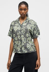 Mladá žena s vrkočmi, oblečená v krátkemu tričku na gombíky so sivým károvaným a žltým kvetinovým vzorom, v kombinácii s čiernymi nohavicami.