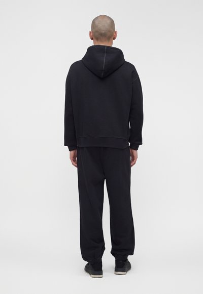 Άτομο που στέκεται με την πλάτη προς τα εμπρός, φοράει μαύρο φούτερ με κουκούλα, μαύρη φόρμα και μαύρα αθλητικά παπούτσια με λευκές σόλες.