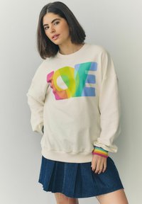 Krémová mikina s nápisom "LOVE" v dúhových farbách, skombinovaná s námorníckou džínovou skladanou sukňou. Rukávy ukazujú viacfarebné pruhy.
