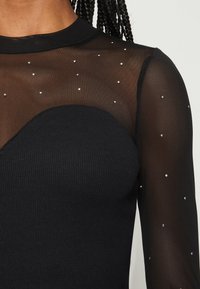 Svart långärmad topp med en genomskinlig meshövre del som har spridda rhinestones och en ribbad del i fast svart nedre sektion.