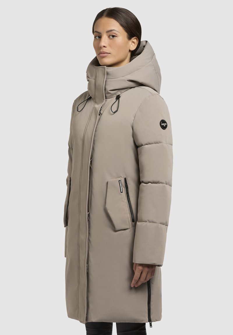 Beige Steppjacke mit hohem Kragen, Frontreißverschluss, Toggle-Akzenten und seitlichen Reißverschlusstaschen. Glatter, strukturierter Stoff mit moderner Silhouette.