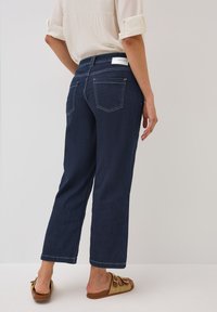 Jeans a vita alta di un blu scuro, con gamba dritta, dotati di due tasche posteriori e cuciture bianche. Abbinati a sandali infradito intrecciati marroni.