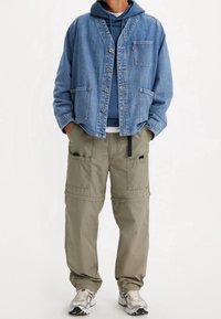 Denim jack met knopen en klepzakken, gedragen over een blauwe hoodie; gecombineerd met loszittende olijfgroene cargobroek en grijze sneakers.