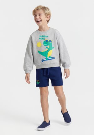 Smiling boy in gray sweatshirt with dinosaur graphic and "Summer Time" text, navy shorts, and matching slip-on shoes walking forward.