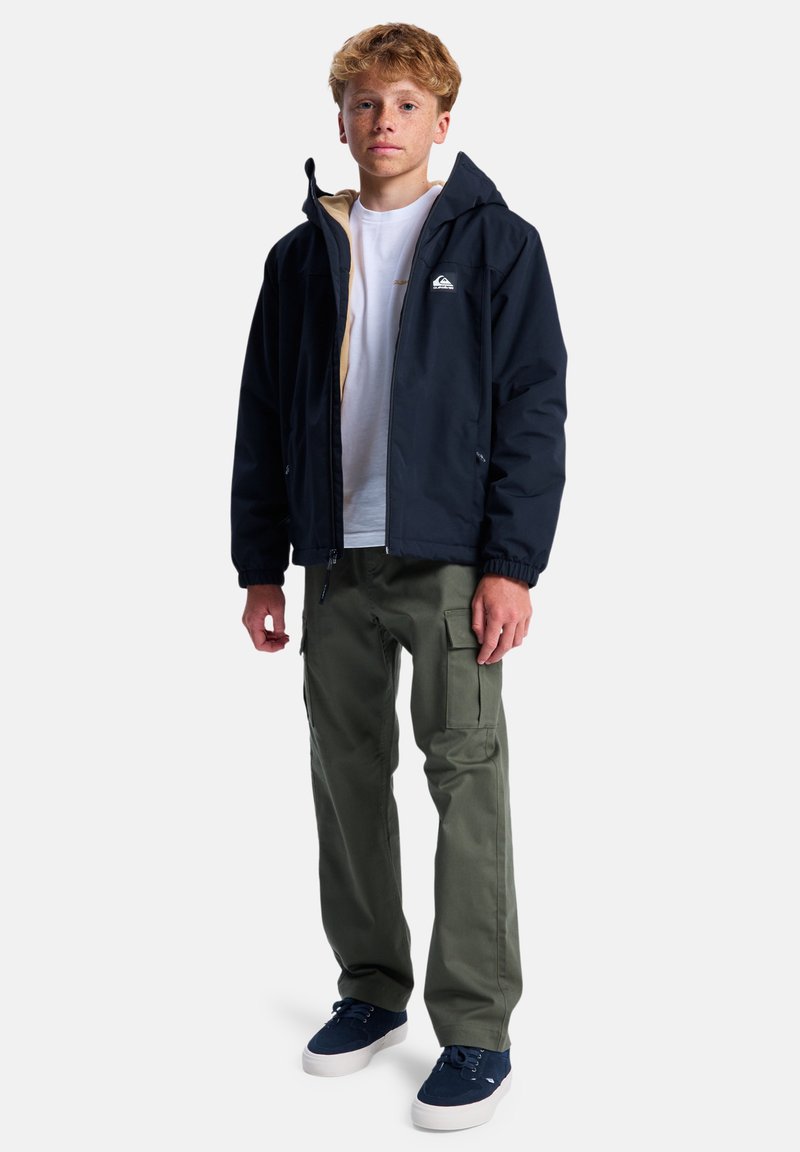 Marineblaue wasserdichte Jacke mit Kapuze, beigem Futter, Reißverschluss vorne; olivgrüne Cargo-Hosen mit Seitentaschen; marineblaue Sneakers mit weißen Sohlen.
